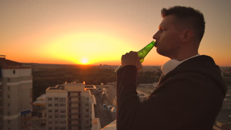 A-man-drinks-beer-standing-on-the-roof-and-contemplates-the-world.-Look-at-the-view-of-the-city-at-sunset-from-a-height.-The-roof-of-a-skyscraper.